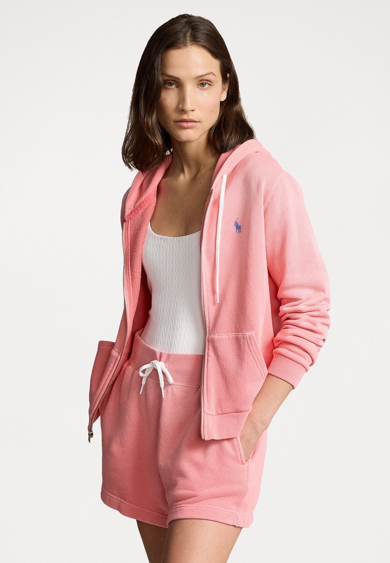 Hoodie de algodão rosa com fecho na frente, calções brancos com cordão e um top de alças branco texturizado. Apresenta um pequeno logo azul no peito.
