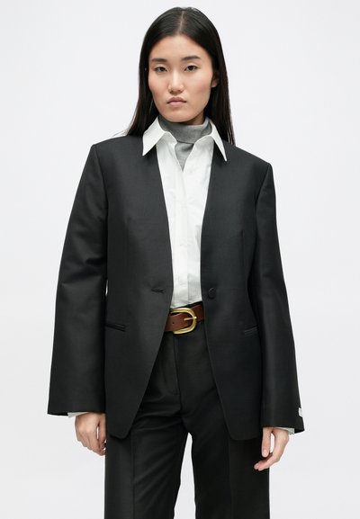 Μαύρη εφαρμοστή σακάκι από λείο υλικό, με ένα κουμπί, πλαϊνές τσέπες και ένα αντίθετο λευκό πουκάμισο από κάτω. Καφέ ζώνη με χρυσά μεταλλικά στοιχεία.