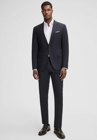 Dunkelblauer Herrenanzug mit maßgeschneiderter Passform, kombiniert mit einem weißen Hemd. Ausgestattet mit einem Reverskragen, Zwei-Knopf-Verschluss und eleganten braunen Schuhen.