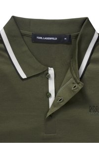 Olivgrön polo med en vit och svart randig krage, tvåknapplikation och broderad logotyp på bröstet. Mjuk, texturerad tyg.