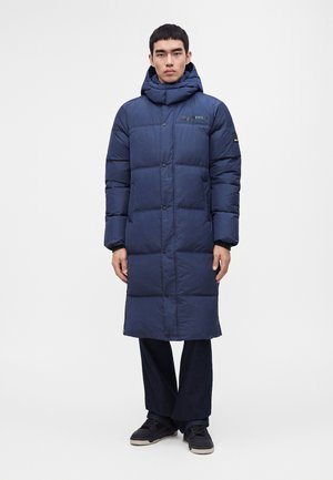 Homme debout portant un long manteau d’hiver matelassé bleu marine avec un col montant et une capuche, associé à un pantalon foncé et des baskets noires.
