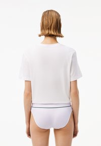 T-shirt en coton blanc coupé avec des manches courtes. Sous-vêtements blancs visibles avec une bande d'accent verte à la taille. Fond uni.