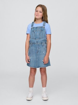 Vestido de sarja de ganga com bainha desfiada, cor lavagem clara, bolso frontal e alças ajustáveis. Combinado com uma camisa azul clara e ténis brancos.