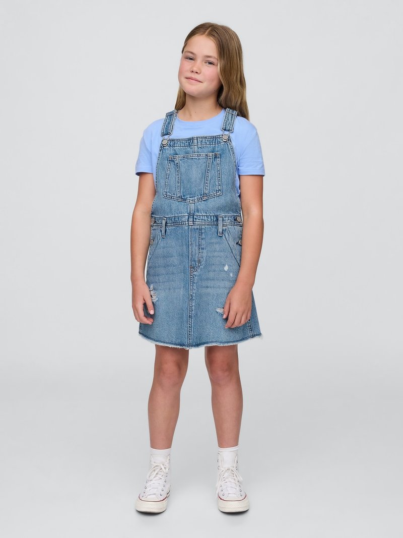 Denim overallkjole med en frynset kant, lys vaskefarge, frontlomme og justerbare stropper. Kombinert med en lyseblå skjorte og hvite sneakers.