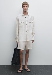 Linen button-up shirt with two chest pockets, paired with matching shorts. Model holds a dark fabric tote bag and wears flip-flops.