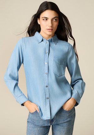 Frau mit langem dunklem Haar, die ein hellblaues Jeanshemd und passende Jeans trägt, die Hände in den Taschen, schaut zur Seite.