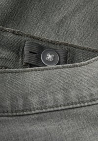 Grå denimtyg med en synlig söm, en transparent knapp och en sydd flik. Texturerad yta med en jämn färg.