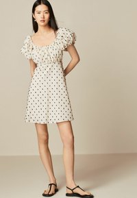 Kvinna med långt mörkt hår som bär en ljusbeige kort klänning med svarta prickar, puffärmar och svarta platta sandaler, stående mot en enkel bakgrund.
