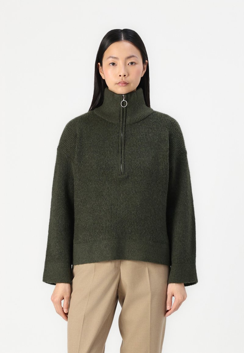 Dunkelgrüner Strickpullover mit hohem Kragen, halbem Reißverschluss vorne und weiten Ärmeln. Strukturierter Stoff und lässige Passform. Kombiniert mit beigen Hosen.