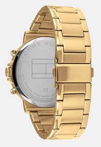 Montre en acier inoxydable doré avec un bracelet à maillons. Elle présente un boîtier rond, un fermoir à bouton-poussoir et un fond gravé avec des détails de logo.