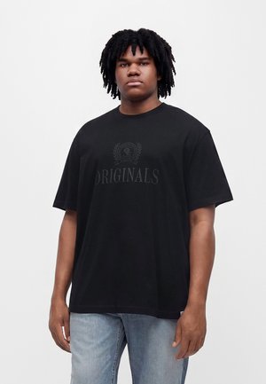 Młody mężczyzna z czarnymi kręconymi włosami, ubrany w czarny t-shirt z napisem "ORIGINALS" oraz jasnoniebieskie dżinsy, stojący na jednolitym białym tle.