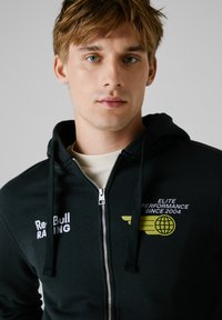 Crni dukserica s patentnim zatvaračem napravljena od pamuka. Sadrži bijeli logo "Red Bull Racing" i žuti grafički tekst na lijevoj strani prsiju. Ima uže za zatezanje s metalnim vrhovima.