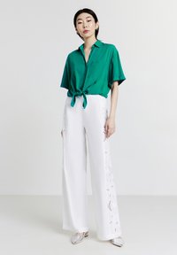 Donna con una camicia verde a maniche corte abbottonata e annodata in vita, pantaloni bianchi a gamba larga con pannelli laterali in pizzo e scarpe argentate a punta.