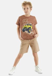 Bruine katoenen t-shirt met een gele jeep grafiek; beige shorts; witte hoge sneakers. Model poseert met de handen in de zakken.