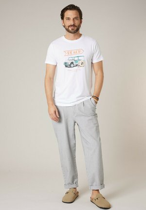 Homme portant un t-shirt blanc avec un graphique de plage, un pantalon léger à rayures, des chaussures beige sans lacets, debout avec une main dans la poche sur un fond neutre.