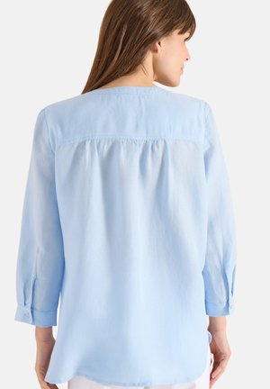 Femme aux cheveux longs et bruns portant un chemisier ample à manches longues bleu clair, vue de dos sur un fond blanc.