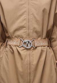 Ceinture beige avec une boucle circulaire en argent arborant les lettres "N" et "V", mettant en avant un tissu texturé et des accents de marque.