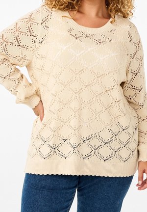 Pull en maille beige avec un motif ajouré en forme de losange, porté avec un jean bleu, montré sur une femme avec une main sur la hanche.