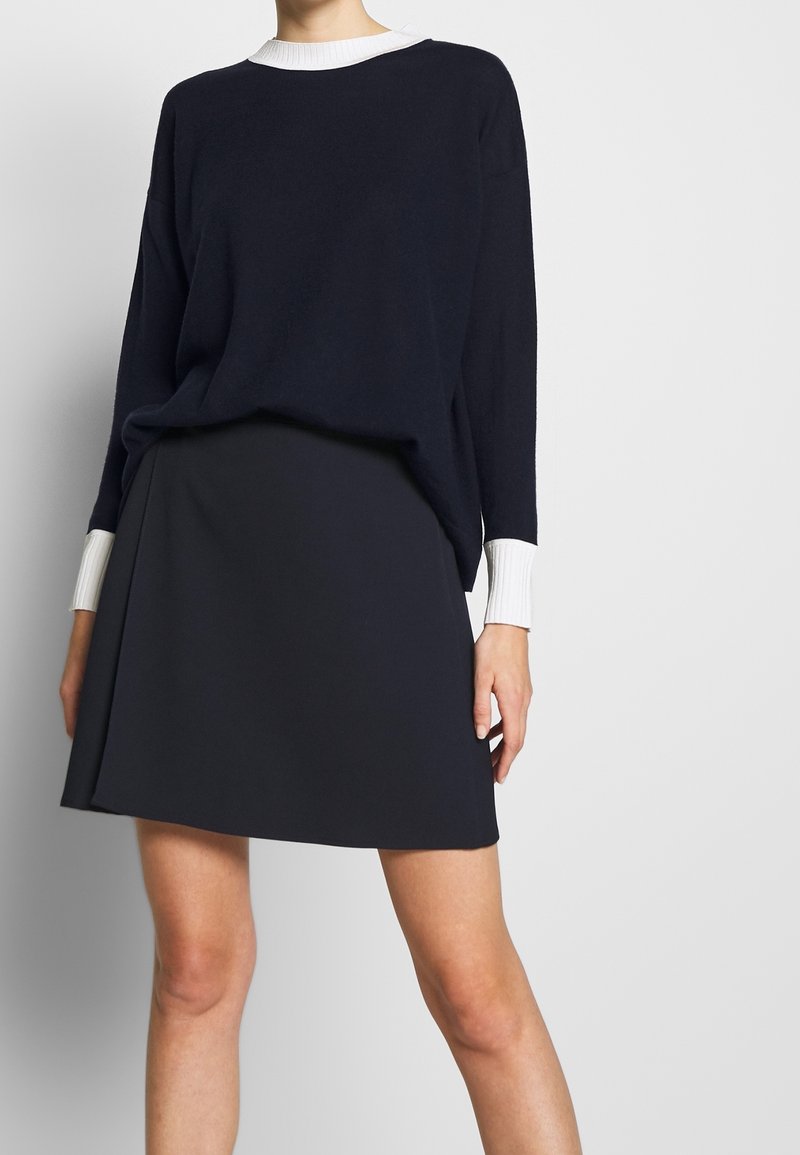 Femme portant un pull noir à manches longues avec des poignets et un col blancs, associé à une mini-jupe noire, sur un fond uni.