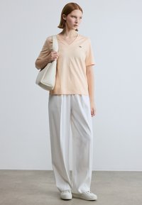 Μπλουζάκι σε απαλό ύφασμα με ντεκολτέ σε σχήμα V σε χρώμα ροδάκινο, συνδυασμένο με άνετο λευκό παντελόνι. Το μοντέλο κρατά μια κρεμ τσάντα χειρός. Αθλητικά λευκά παπούτσια ολοκληρώνουν την εμφάνιση.