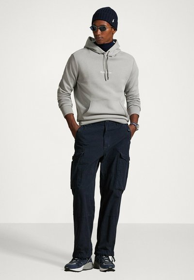 Felpa con cappuccio grigio chiaro con tasca frontale, abbinata a pantaloni cargo blu navy con tasche laterali. Accessori includono un berretto scuro e occhiali da sole.
