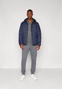 Mörkblå vadderad jacka med huva, blixtlåsstängning och en logotyp på bröstet, kombinerad med en grå t-shirt med tryck och grå joggingbyxor. Vita sneakers.