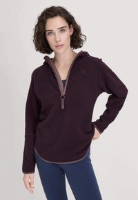 Frau mit kurzen, lockigen Haaren, die einen dunkelvioletten Fleece-Pullover mit halbem Reißverschluss und marineblauen Leggings trägt und vor einem einfachen, hellen Hintergrund steht.