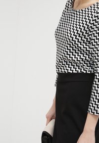 Haut à motif zigzag noir et blanc avec des manches trois quarts, associé à une jupe noire, retenue par une pochette de couleur claire.