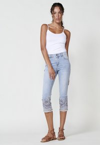 Light blue capri jeans with embroidered accents at the cuffs, featuring a slim fit and a mid-rise waist. Paired with a white tank top and brown sandals.