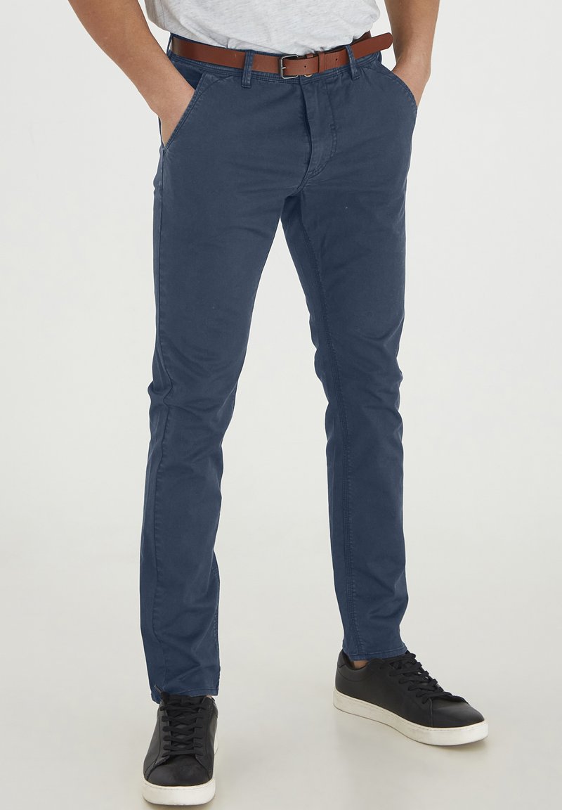 Námornícké modré chinos nohavice, bavlnený materiál, slim fit, dizajn s piatimi vreckami, kombinované s čiernymi teniskami a hnedým koženým opaskom pre kontrast.