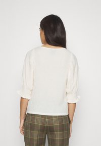 Blusa crema con maniche a sbuffo e polsini arricciati. Tessuto con trama, scollatura rotonda, indossata con pantaloni a quadri, verdi e marroni.