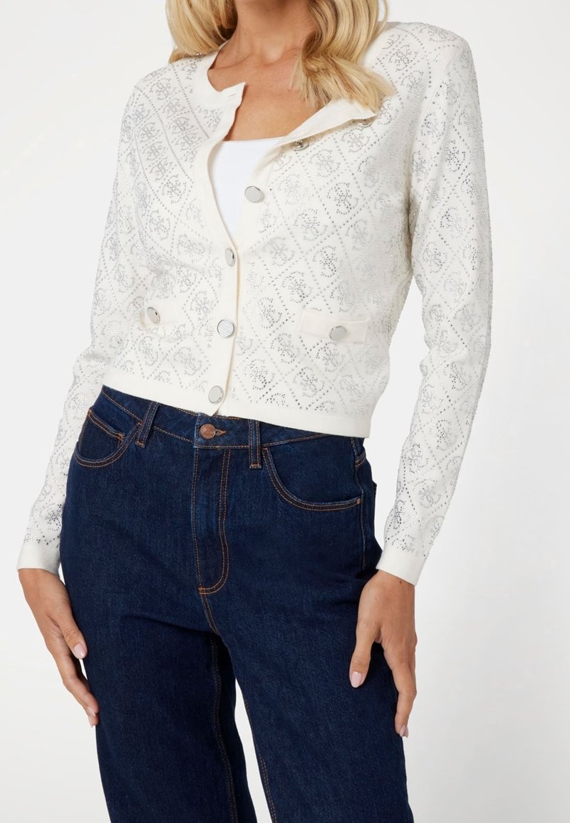 Femme portant un cardigan blanc à motifs boutonné par-dessus un haut blanc, assorti à un jean bleu foncé taille haute sur fond blanc.