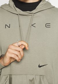 Světle zelená mikina s kapucí vyrobená z měkké látky. Obsahuje černé nápisy "NIKE" a logo. Má přední kapsu s texturovaným logem.