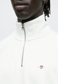 Fehér cipzáras pulóver magas gallérral, ezüst cipzárral és a mellkason egy kis hímzett logóval. Simább anyagtextúra.