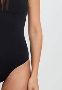 Maillot de bain une pièce noir avec un panneau en mesh transparent au niveau du col. Texture lisse, design ajusté et ouvertures de jambe hautes.