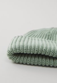 Bonnet en tricot de couleur vert clair, avec un design en côtes repliées. Matériau texturé et doux avec des coutures visibles et une finition lisse.