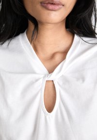 White cotton shirt featuring a knot detail at the neckline and a keyhole cutout below. Smooth texture with a relaxed fit.