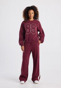 Bordeaux sweatshirt met witte "89" print, gecombineerd met bijpassende wijde broek met witte zijstrepen, gemaakt van zachte stof.