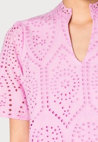 Camicia di cotone rosa con intricati modelli intagliati, colletto alla mandarino e maniche corte. Superficie testurizzata con ricami decorativi.