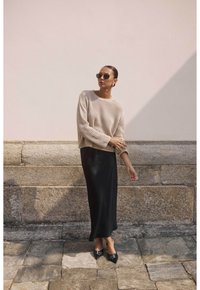 Pull en tricot beige à texture côtelée, associé à une jupe maxi noire ajustée. La mannequin porte des lunettes de soleil sombre et des ballerines noires à bout pointu.