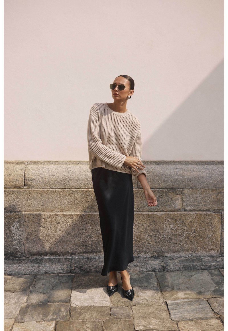 Pull en tricot beige à texture côtelée, associé à une jupe maxi noire ajustée. La mannequin porte des lunettes de soleil sombre et des ballerines noires à bout pointu.