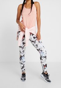 Jasnoróżowy top na ramiączkach związywany na talii, w zestawie z wielokolorowymi legginsami w kwiaty i czarnymi butami sportowymi.