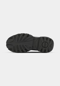 Semelle de chaussure noire avec des rainures profondes, des rainures incurvées pour l’adhérence et un logo circulaire de la marque près du talon sur un fond gris clair.