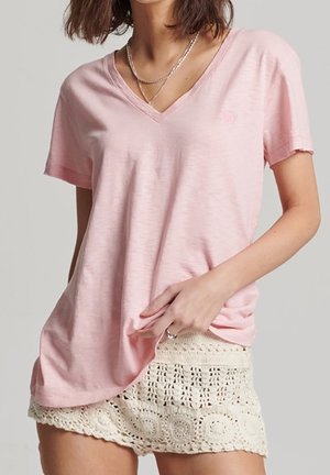 Vrouw draagt een lichtroze V-hals T-shirt met korte mouwen en crèmekleurige gehaakte shorts, gecombineerd met gelaagde kettingen.