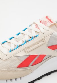 Beige sneakers med mesh- och lädermaterial, med röda och blå accenter, platta vita snören och en strukturerad sula. Reebok-logotyp på sidan.