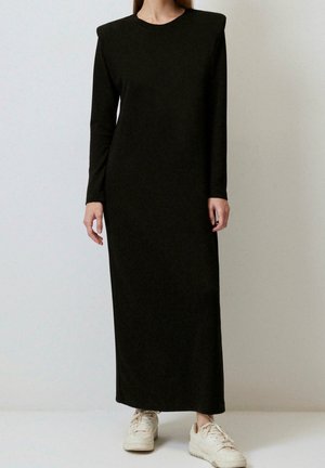 Femme portant une longue robe noire avec des épaules rembourrées et des baskets blanches, debout devant un fond uni.