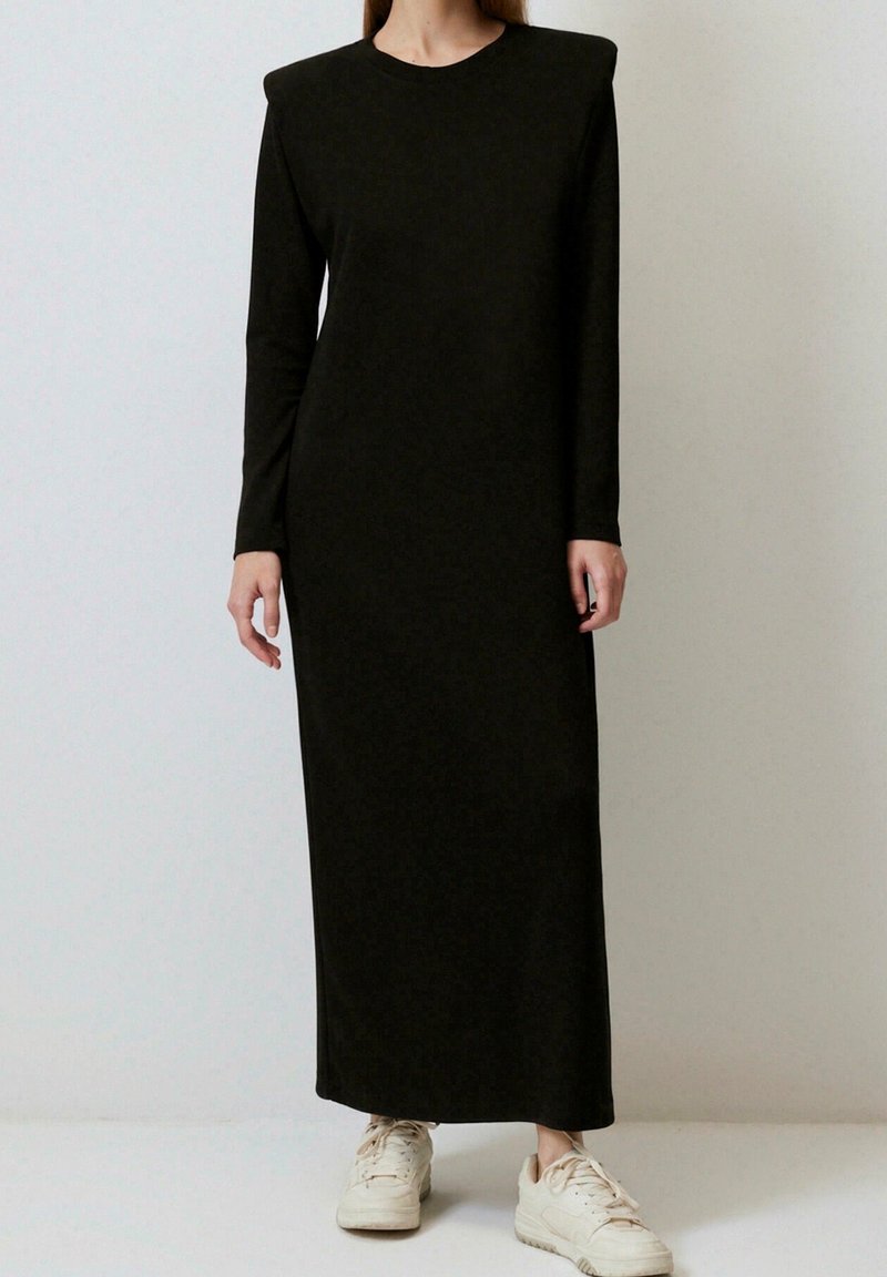 Femme portant une longue robe noire avec des épaules rembourrées et des baskets blanches, debout devant un fond uni.