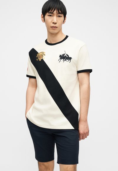 Νέος άνδρας που φοράει λευκό μπλουζάκι με μαύρη διαγώνια ρίγα, λογότυπο παίκτη πόλο και ναυτικό μπλε σορτς, στέκεται με τα χέρια στις τσέπες μπροστά σε απλό φόντο.