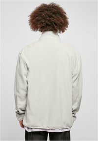 Vaaleanharmaa fleece-takki, jossa on korkea kaulus, pitkät hihat ja ribbireunat. Kiva sileä pinta, jossa on kontrastoivaa kangasta hihoissa.