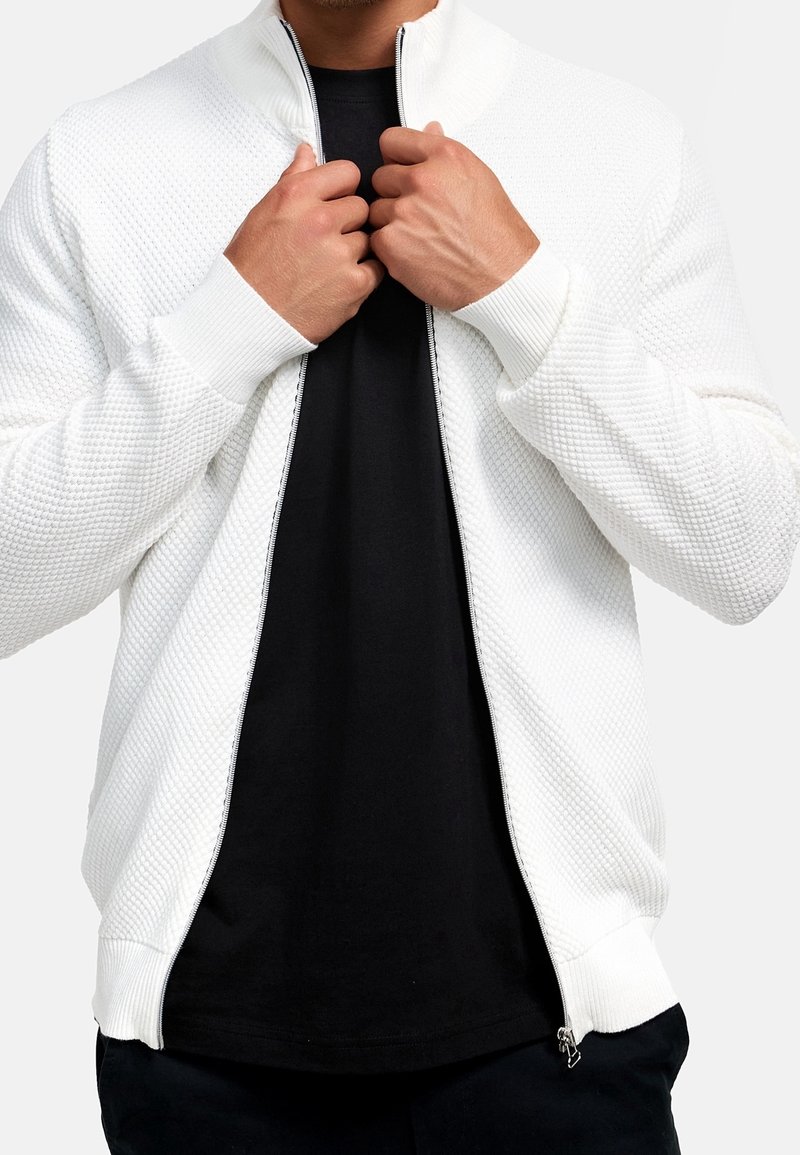 Valkoinen vetoketjullinen takki, jossa on teksturoitu pinta, ribbattuja hihansuita ja kaulus; käytetään mustan paidan päällä. Vetoketju yksityiskohtana näkyvissä edessä.